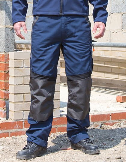 Das Bild zeigt einen Mann, der eine Arbeitshose in Blau mit dunkelblauen Akzenten trägt. Die Hose ist mit Knieschutz ausgestattet, um den Träger vor Verletzungen und Abnutzung zu schützen. Der Mann trägt auch eine passende Jacke und Sicherheitsschuhe, die seine Arbeitskleidung vervollständigen. Die Hose hat mehrere Taschen, um Werkzeuge und andere Arbeitsutensilien sicher aufzubewahren. Der Mann sieht bereit aus, um seine Arbeit sicher und effektiv zu erledigen. 