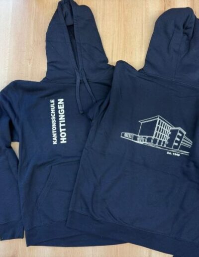 Zwei navyblaue Hoodies mit Textildruck: Schulname vorne, Gebäudeskizze mit "Est. 1976" und Schriftzug auf dem Rücken.