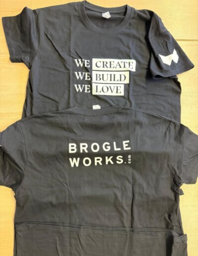 Zwei schwarze T-Shirts auf Holz, bedruckt mit Slogan und Firmenlogo. Textildruck für Firmen- oder Vereinskleidung in der Schweiz.