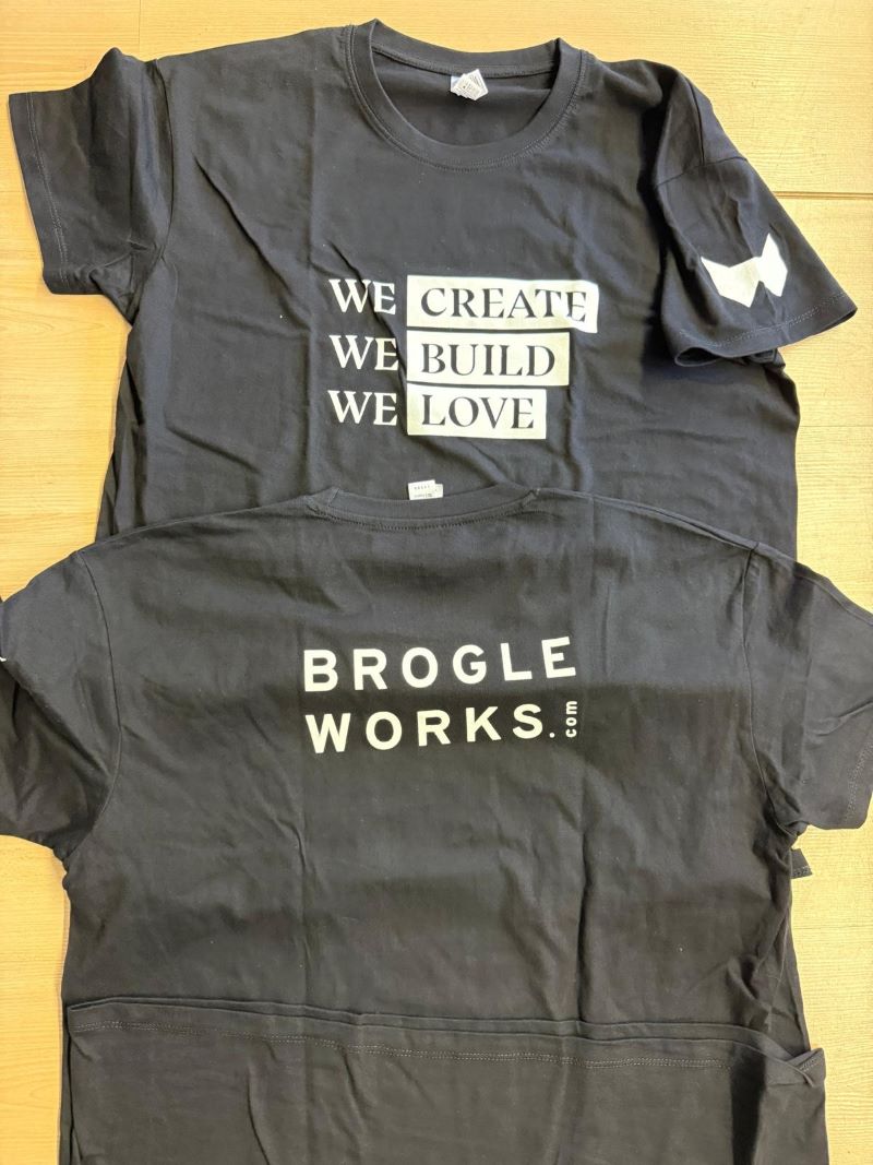 Zwei schwarze T-Shirts auf Holz, bedruckt mit Slogan und Firmenlogo. Textildruck für Firmen- oder Vereinskleidung in der Schweiz.